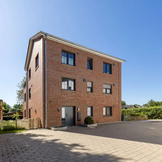 Dreistöckiges Wohnhaus aus Backstein mit mehreren Fenstern, einer flachen, gepflasterten Fläche davor und einem klaren, blauen Himmel über dem Haus. - Bauunternehmen Winsen und Landkreis Harburg