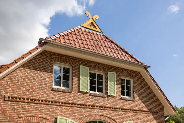 Ein Backsteinhaus mit grünen Fensterläden, drei Fenstern und einem roten Ziegeldach unter einem blauen Himmel mit einigen Wolken. - Bauunternehmen Winsen und Landkreis Harburg