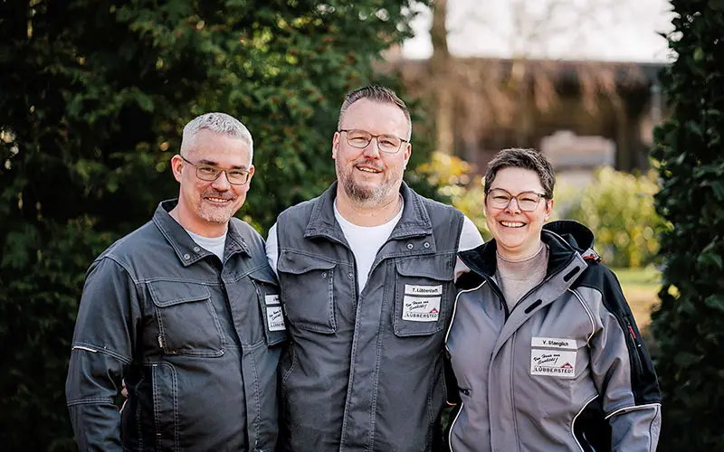 Drei Personen in passenden grauen Arbeitsjacken stehen im Freien, lächeln in die Kamera und sind von Grün umgeben. - Bauunternehmen Winsen und Landkreis Harburg
